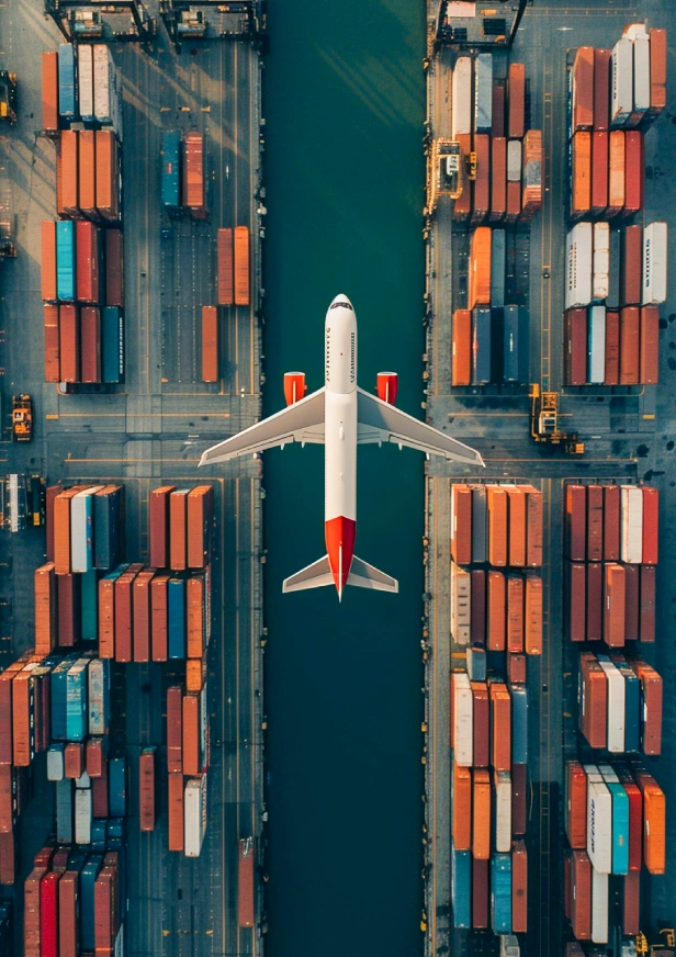 Aerial view of a white and red airplane flying over a canal, surrounded by rows of colorful shipping containers at a port terminal, highlighting Air Freight Shipping Services in Dubai.