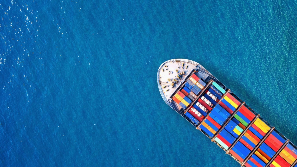 Aerial view of a cargo ship with colorful shipping containers, highlighting ocean freight shipping, on bright blue water, with the ship positioned diagonally in the lower right corner.