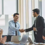 Two businessmen in suits shake hands and smile during a meeting at a modern Shipping Company Dubai office, while three colleagues sit at a table, clapping and showing support. Laptops and documents are visible on the table.