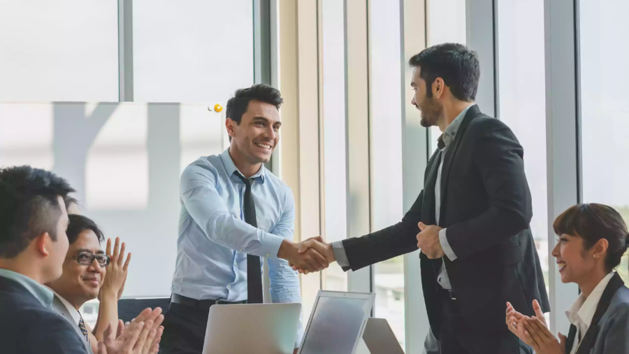 Two businessmen in suits shake hands and smile during a meeting at a modern Shipping Company Dubai office, while three colleagues sit at a table, clapping and showing support. Laptops and documents are visible on the table.