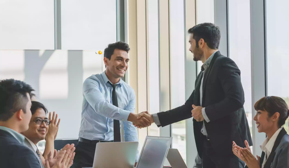 Two businessmen in suits shake hands and smile during a meeting at a modern Shipping Company Dubai office, while three colleagues sit at a table, clapping and showing support. Laptops and documents are visible on the table.
