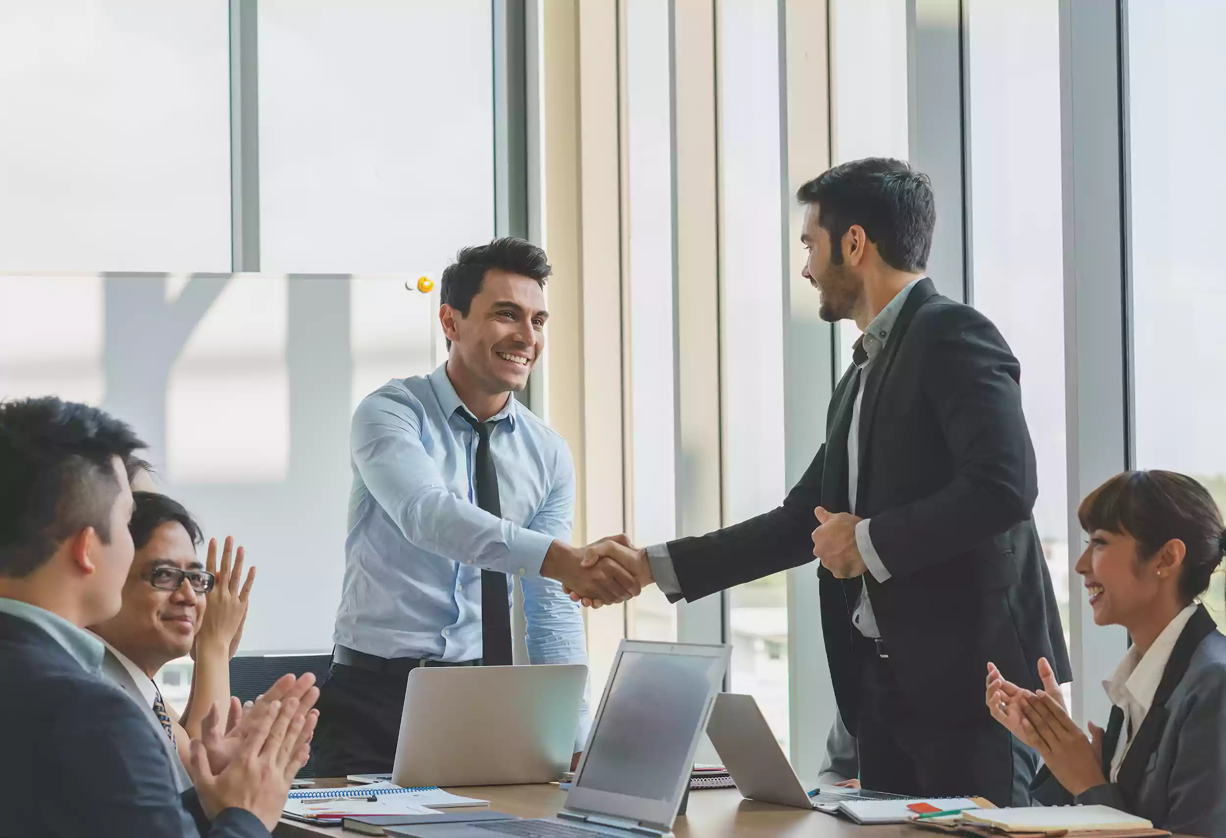 Two businessmen in suits shake hands and smile during a meeting at a modern Shipping Company Dubai office, while three colleagues sit at a table, clapping and showing support. Laptops and documents are visible on the table.