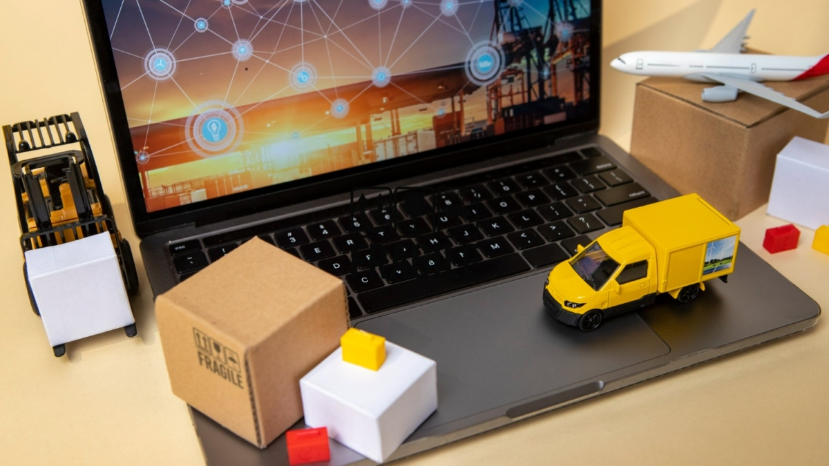 Laptop displaying a global shipping network graphic, alongside miniature forklift, delivery truck, and various cardboard boxes, illustrating e-commerce logistics.