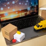 Laptop displaying a global shipping network graphic, alongside miniature forklift, delivery truck, and various cardboard boxes, illustrating e-commerce logistics.