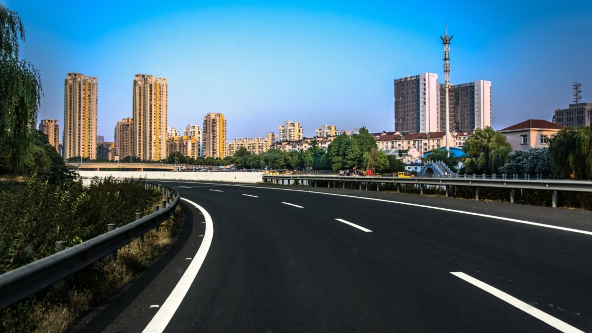 A wide, empty asphalt road—part of the impressive UAE road infrastructure—curves toward a city skyline with tall, modern buildings under a clear blue sky, bordered by trees and greenery on both sides.