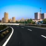 A wide, empty asphalt road—part of the impressive UAE road infrastructure—curves toward a city skyline with tall, modern buildings under a clear blue sky, bordered by trees and greenery on both sides.